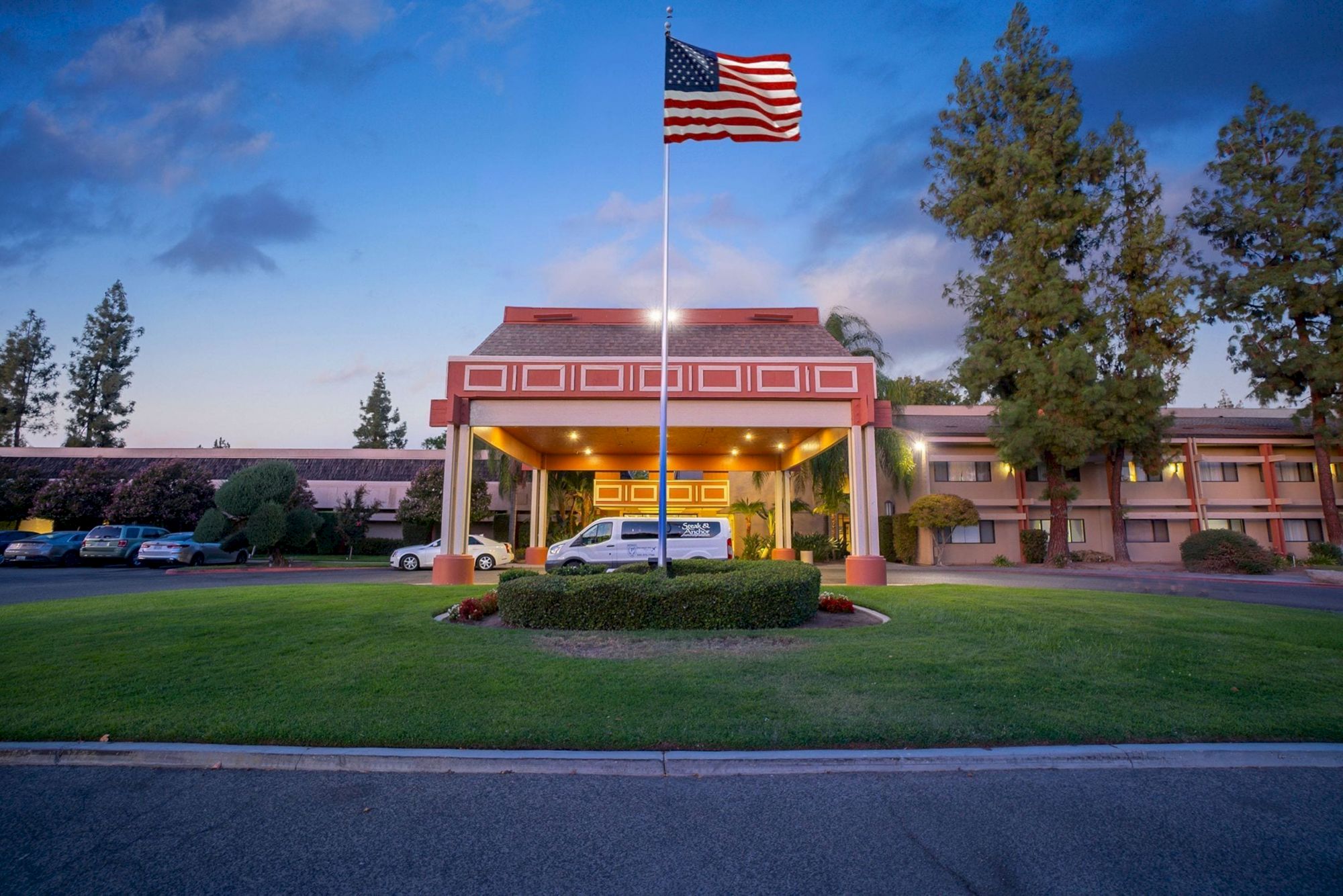 A building with a prominent entrance and flagpole, displaying the US flag, surrounded by trees and a grassy area, under a twilight sky.