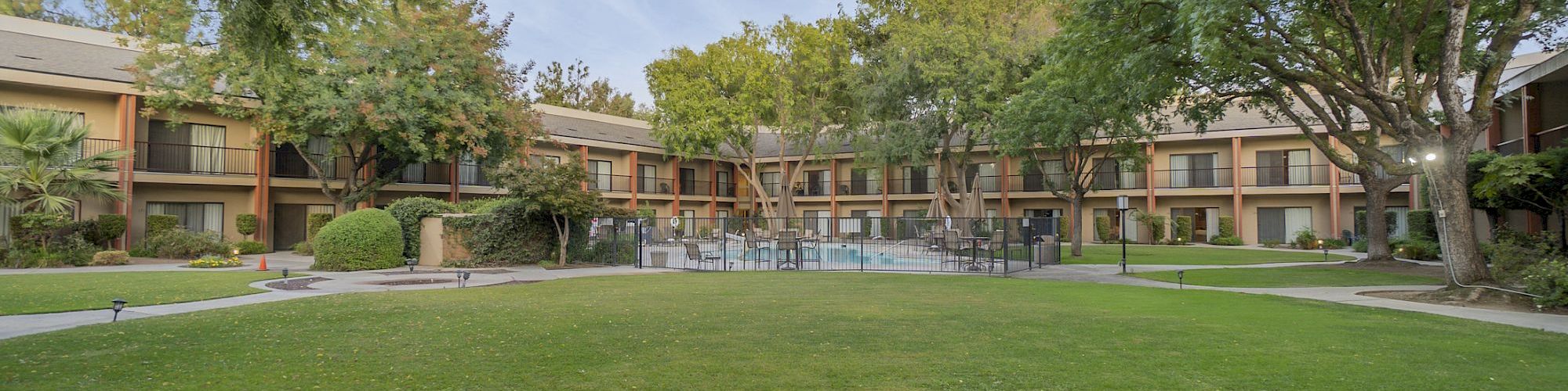 A large manicured lawn is surrounded by a two-story building with balconies, trees, and a central pool area under a clear blue sky.